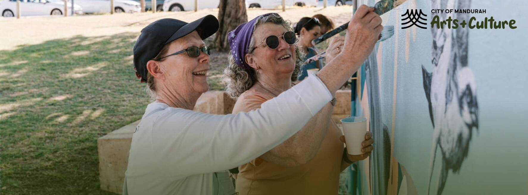 Two people smiling while painting a blue mural together at an outdoor community art event